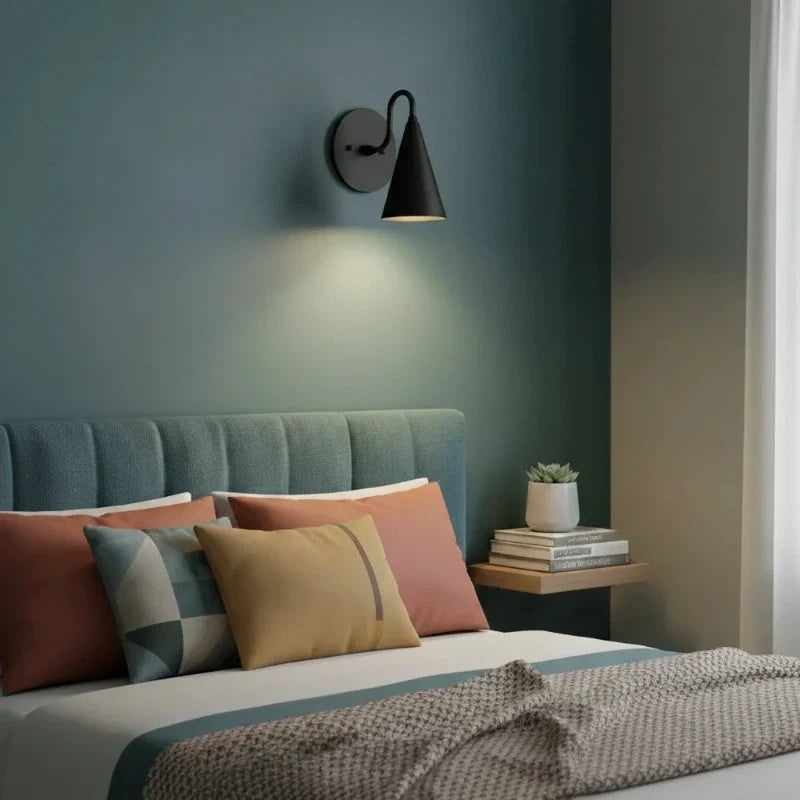 Bedroom with a bed featuring colorful pillows and a nightstand with books and a plant, illuminated by a wall lamp.